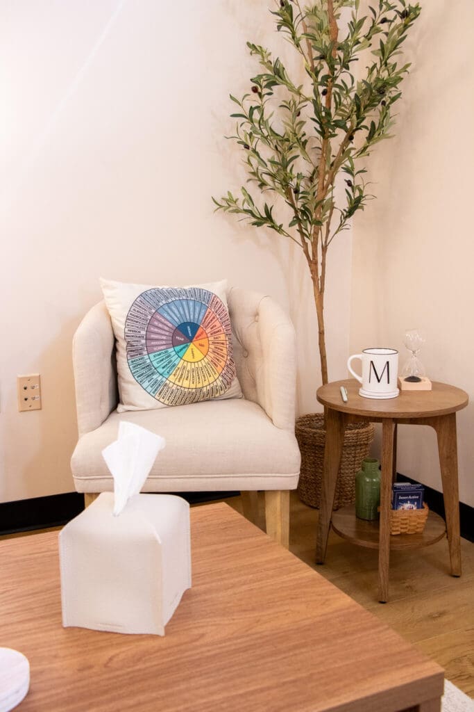 An emotion wheel pillow rests on a cozy chair beside a plant and side table. What does trauma feel like when it's finally given a name? Christian counseling in Gilbert, AZ uses tools like this to help clients process it safely.